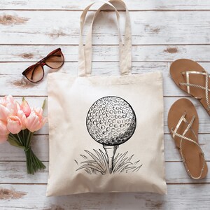 May include: A white canvas tote bag with a black and white illustration of a golf ball on a tee. The golf ball is in the center of the bag and is surrounded by grass.
