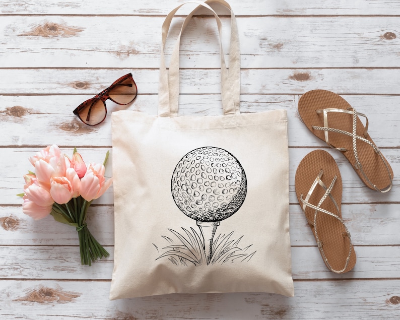 May include: A white canvas tote bag with a black and white illustration of a golf ball on a tee. The golf ball is in the center of the bag and is surrounded by grass.