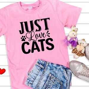 Może przedstawiać: R&oacute;żowa koszulka z napisem "Just Love Cats" czarnymi literami. Koszulka leży na białym drewnianym tle z niebieskimi dżinsami, dwoma czerwonymi sercami i dwoma pluszowymi misiami.
