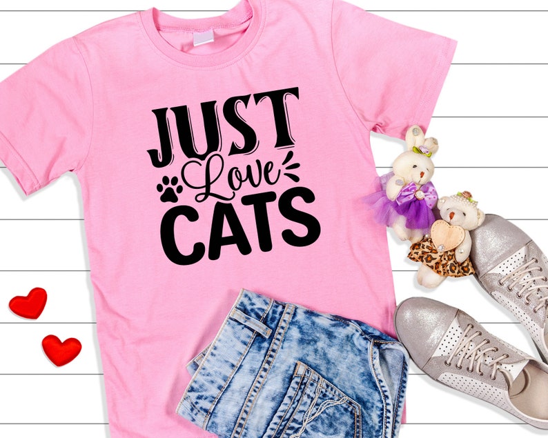 Może przedstawiać: R&oacute;żowa koszulka z napisem "Just Love Cats" czarnymi literami. Koszulka leży na białym drewnianym tle z niebieskimi dżinsami, dwoma czerwonymi sercami i dwoma pluszowymi misiami.