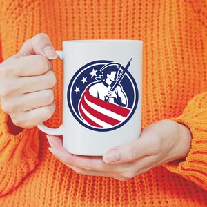 K&ouml;nnte beinhalten: Wei&szlig;e Keramiktasse mit einer Grafik einer Person in einem Dreispitzhut, die ein Gewehr h&auml;lt und vor einer amerikanischen Flagge steht. Die Grafik befindet sich in einem Kreis mit drei Sternen &uuml;ber der Person.