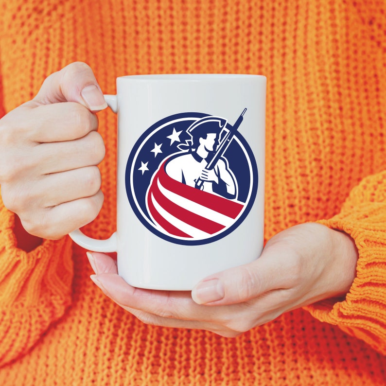 K&ouml;nnte beinhalten: Wei&szlig;e Keramiktasse mit einer Grafik einer Person in einem Dreispitzhut, die ein Gewehr h&auml;lt und vor einer amerikanischen Flagge steht. Die Grafik befindet sich in einem Kreis mit drei Sternen &uuml;ber der Person.