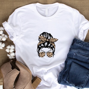 Op de afbeelding: Wit T-shirt met een afbeelding van een vrouw met een rommelige knot, een luipaardprint hoofdband en een zonnebril.