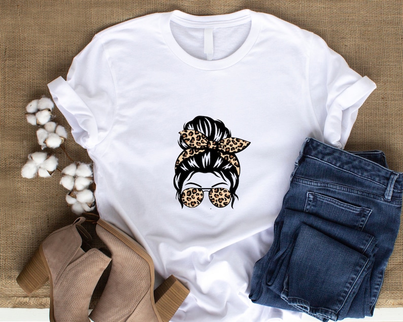Op de afbeelding: Wit T-shirt met een afbeelding van een vrouw met een rommelige knot, een luipaardprint hoofdband en een zonnebril.