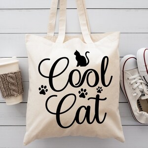 Może przedstawiać: Biała pł&oacute;cienna torba na zakupy z czarnym napisem "Cool Cat" i sylwetką kota. Na torbie znajdują się trzy odciski łap.