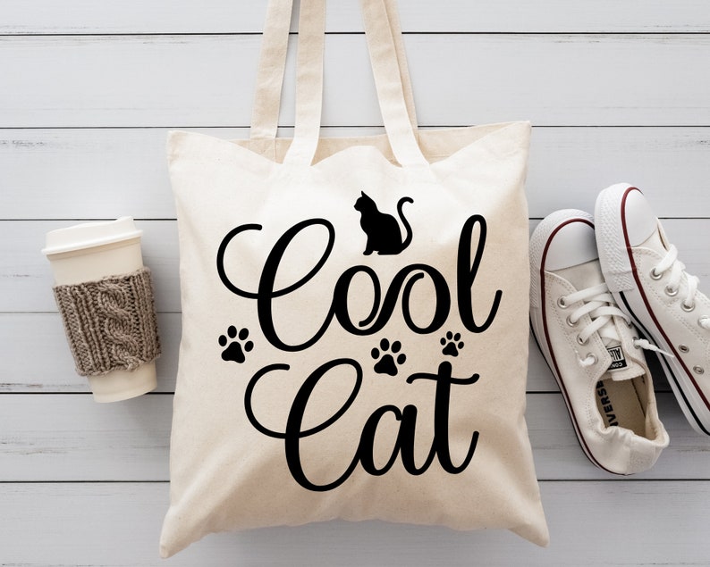 Może przedstawiać: Biała pł&oacute;cienna torba na zakupy z czarnym napisem "Cool Cat" i sylwetką kota. Na torbie znajdują się trzy odciski łap.