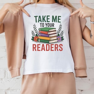 以下が含まれることがあります： 緑と赤で「TAKE ME TO YOUR READERS」と書かれた白いTシャツ。緑、赤、紫、黄色の本が積み重ねられ、花のアクセントが付いています。カジュアルで快適なTシャツです。