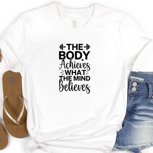 Op de afbeelding: Wit T-shirt met zwarte tekst die "The Body Achieves What The Mind Believes" luidt, met een halterstangsymbool aan beide kanten van het woord "The".