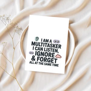 K&ouml;nnte beinhalten: Eine wei&szlig;e Karte mit dem Text "I AM A MULTITASKER I CAN LISTEN, IGNORE & FORGET ALL AT THE SAME TIME" in Schwarz, T&uuml;rkis und Lila. Die Karte liegt auf einem wei&szlig;en Teller und einem beigen Stoff.