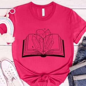 May include: A pink t-shirt with a black graphic of an open book with crystals and stars inside.