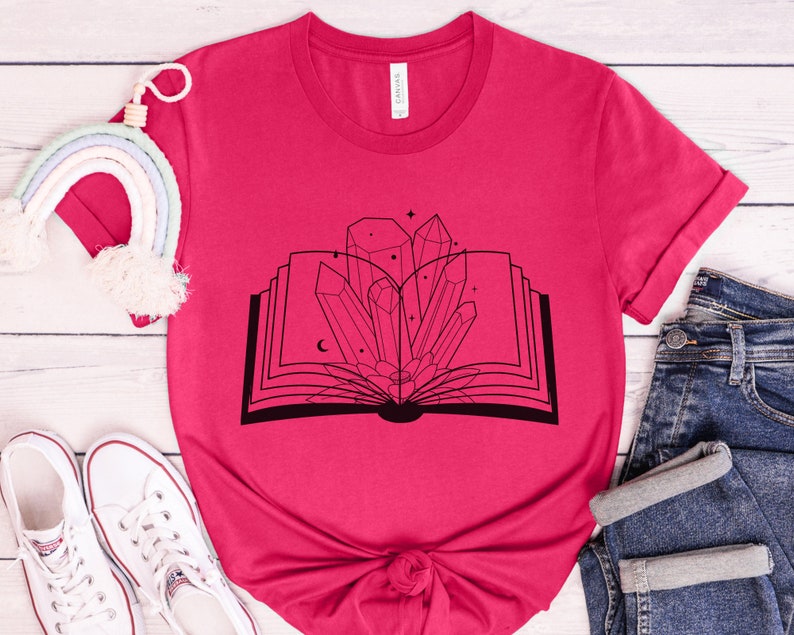 May include: A pink t-shirt with a black graphic of an open book with crystals and stars inside.