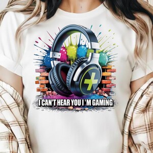 May include: White t-shirt featuring a graphic of black and silver headphones with a green plus sign on the ear cup. The design includes colorful paint splatters and a brick wall background. Text reads: "I CAN'T HEAR YOU I'M GAMING."