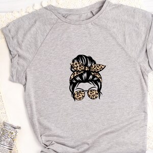 Op de afbeelding: Een grijs T-shirt met een afbeelding van een vrouw met een rommelige knot, een luipaardprint hoofdband en een zonnebril.