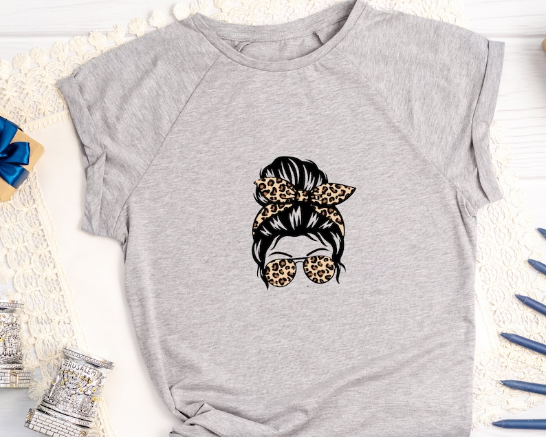 Op de afbeelding: Een grijs T-shirt met een afbeelding van een vrouw met een rommelige knot, een luipaardprint hoofdband en een zonnebril.