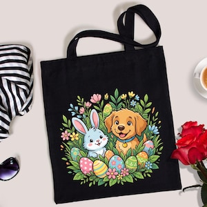 May include: A black tote bag featuring a colorful graphic of a light blue rabbit and a golden puppy surrounded by green foliage and patterned Easter eggs. The design is centered on the front of the bag, which has two black fabric shoulder straps.
