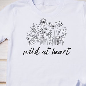 Puede incluir: Camiseta blanca con un dibujo de l&iacute;nea negra de flores silvestres y el texto "wild at heart".