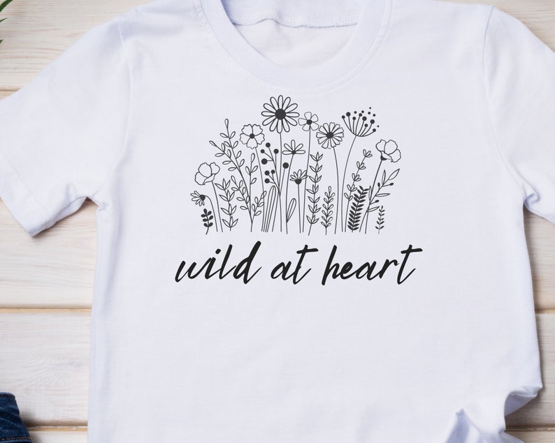 Puede incluir: Camiseta blanca con un dibujo de l&iacute;nea negra de flores silvestres y el texto "wild at heart".