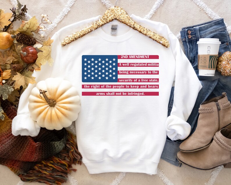 以下が含まれることがあります： 赤、白、青のアメリカ国旗のグラフィックと「2ND AMENDMENT」のテキスト、および「A well regulated militia being necessary to the security of a free state, the right of the people to keep and bear arms shall not be infringed.」のテキストが書かれた白いスウェットシャツ