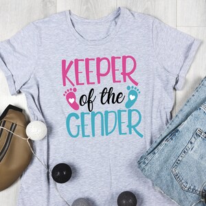 May include: Grey t-shirt with the text "KEEPER of the GENDER" in pink and blue lettering. The text is surrounded by two blue and pink footprints with white hearts.