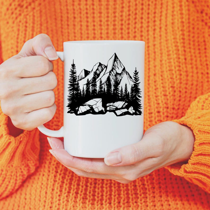 May include: White ceramic mug with a black line drawing of a mountain range and trees. The mug is being held by a person wearing an orange sweater.