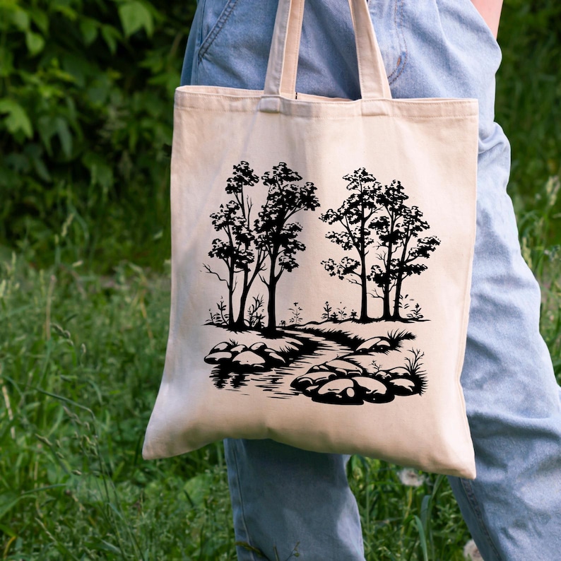May include: A beige tote bag with black silhouette artwork of trees and a stream. The artwork features a winding stream with rocks and foliage.
