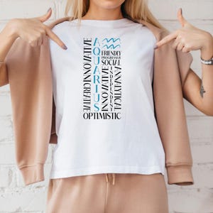 May include: White t-shirt with the word "AQUARIUS" in a vertical design, with related words like "innovative" and "optimistic". The Aquarius symbol is at the top. The shirt is worn with a tan jacket and pants.