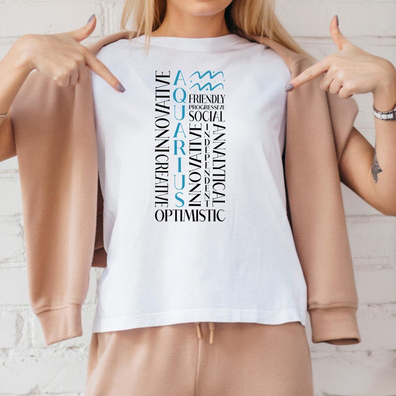 May include: White t-shirt with the word "AQUARIUS" in a vertical design, with related words like "innovative" and "optimistic". The Aquarius symbol is at the top. The shirt is worn with a tan jacket and pants.