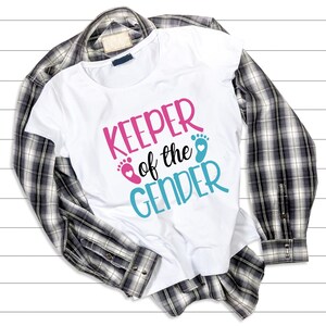 May include: White t-shirt with the text "KEEPER of the GENDER" in pink and blue lettering with a pink and blue footprint graphic. The shirt is folded and laid on a plaid shirt.