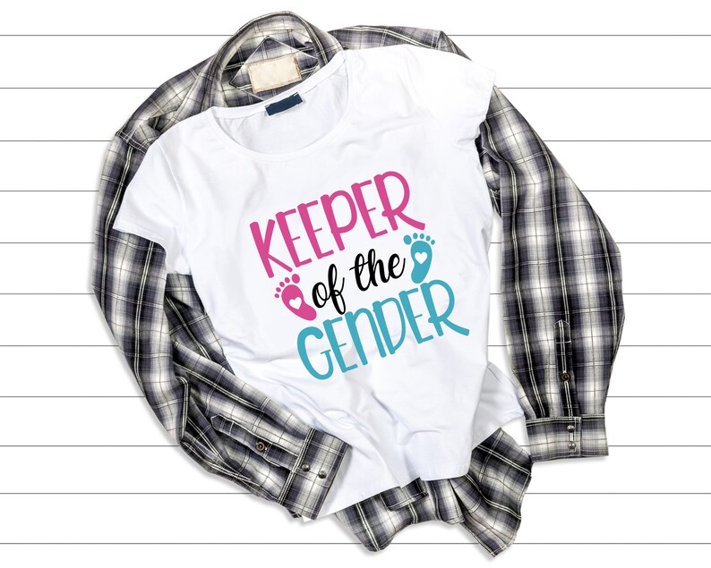 May include: White t-shirt with the text "KEEPER of the GENDER" in pink and blue lettering with a pink and blue footprint graphic. The shirt is folded and laid on a plaid shirt.