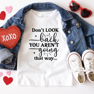 May include: White t-shirt with black text that reads "Don't look back you aren't going that way..."  The shirt is laid out on a white surface with a denim jacket, red heart-shaped sunglasses, and red and pink felt hearts.