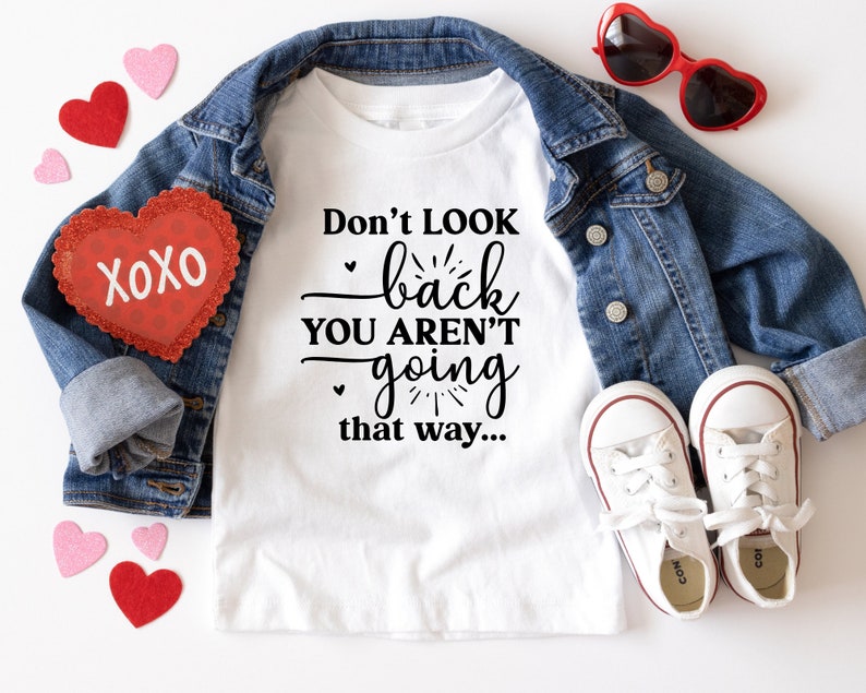 May include: White t-shirt with black text that reads "Don't look back you aren't going that way..."  The shirt is laid out on a white surface with a denim jacket, red heart-shaped sunglasses, and red and pink felt hearts.