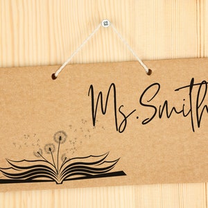May include: A brown cardboard sign with the text "Ms. Smith" in black cursive writing. The sign has a black and white illustration of an open book with dandelions blowing in the wind.