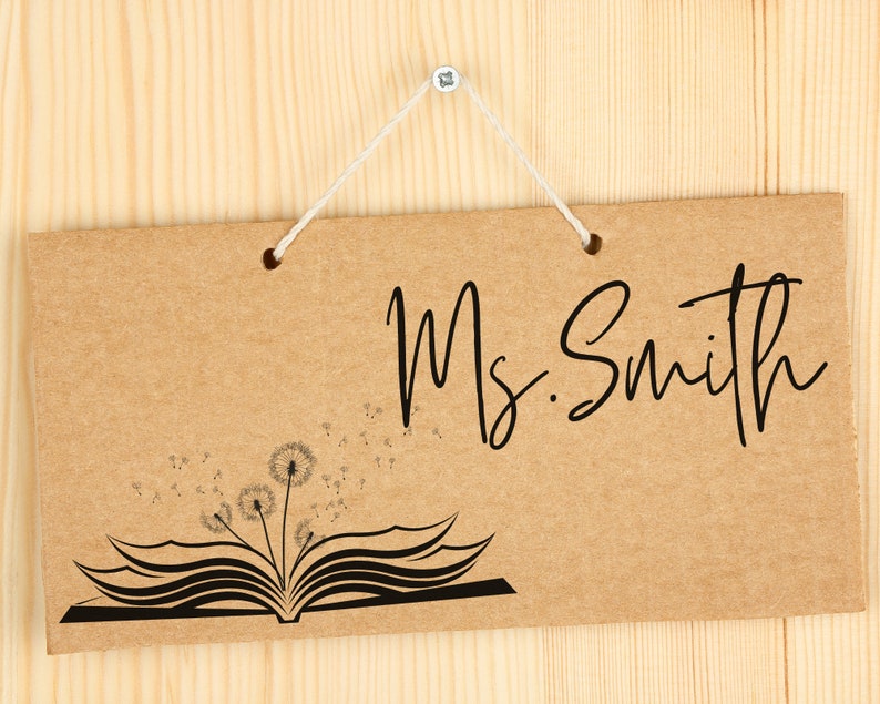 May include: A brown cardboard sign with the text "Ms. Smith" in black cursive writing. The sign has a black and white illustration of an open book with dandelions blowing in the wind.