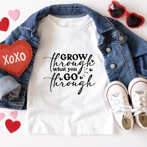 May include: White t-shirt with the text "GROW through what you GO through" in black script with small hearts. The shirt is laid out on a white surface with a denim jacket, red heart-shaped sunglasses, and red and pink heart-shaped decorations.