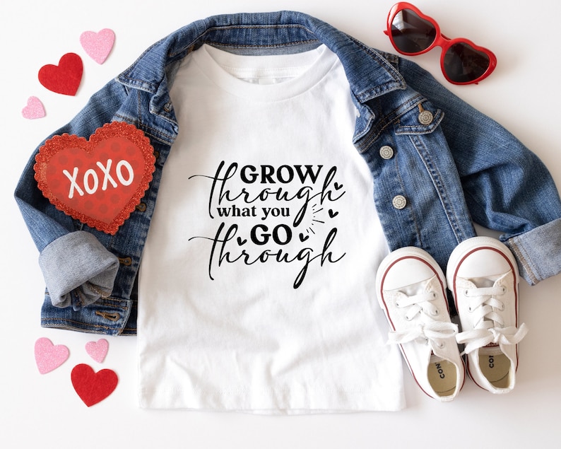 May include: White t-shirt with the text "GROW through what you GO through" in black script with small hearts. The shirt is laid out on a white surface with a denim jacket, red heart-shaped sunglasses, and red and pink heart-shaped decorations.