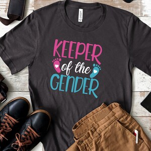 May include: A dark grey t-shirt with the text "Keeper of the Gender" in pink and blue lettering. The text is accompanied by a pink footprint with a white heart and a blue footprint with a white heart.