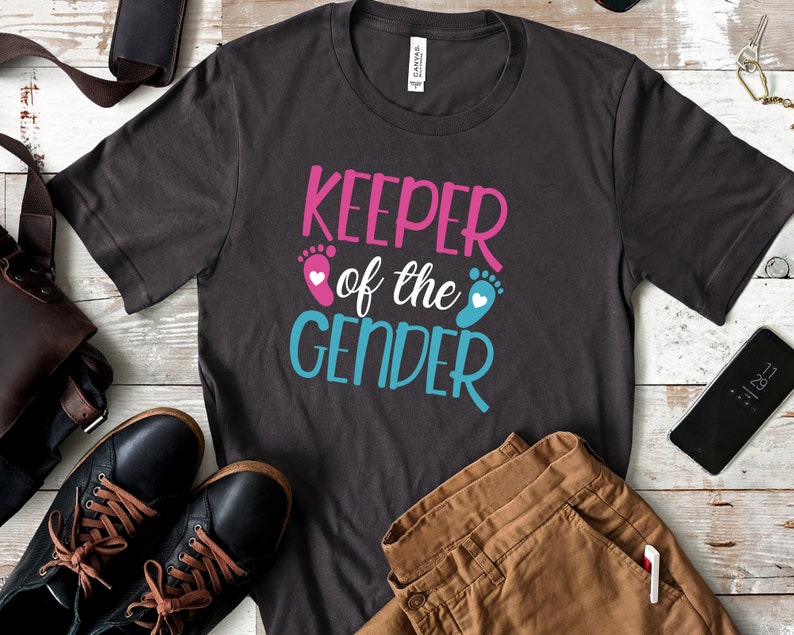 May include: A dark grey t-shirt with the text "Keeper of the Gender" in pink and blue lettering. The text is accompanied by a pink footprint with a white heart and a blue footprint with a white heart.