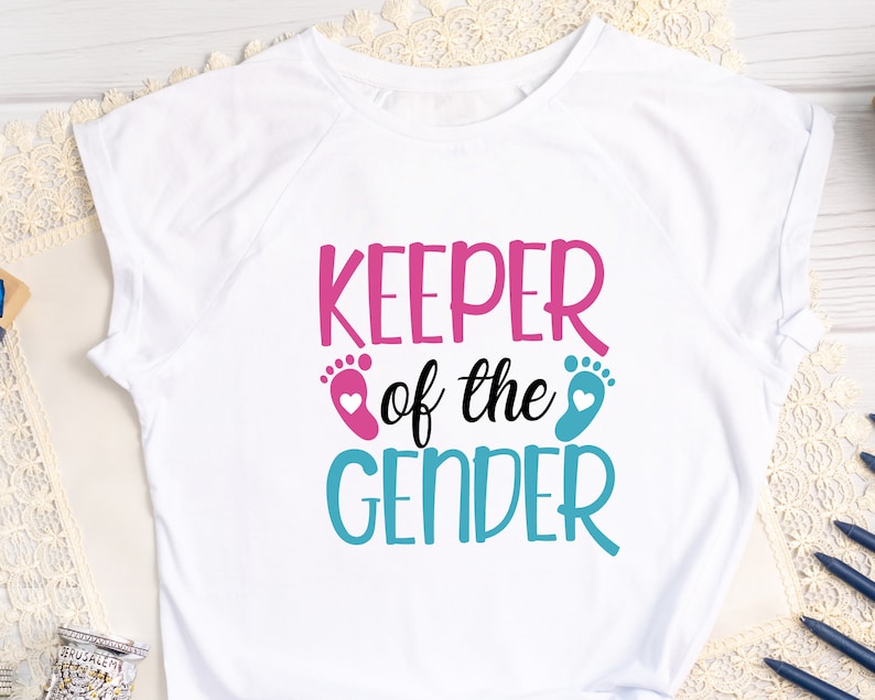 May include: White t-shirt with the text "KEEPER of the GENDER" in pink and blue lettering. The text is accompanied by a pink and blue footprint with a heart.