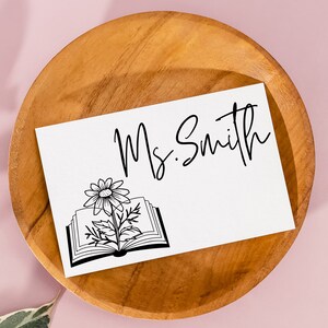 May include: A white card with black text that reads "Ms. Smith" and a black and white illustration of a book with a flower growing out of it. The card is sitting on a wooden plate.