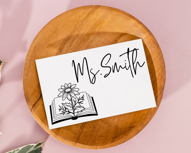 May include: A white card with black text that reads "Ms. Smith" and a black and white illustration of a book with a flower growing out of it. The card is sitting on a wooden plate.