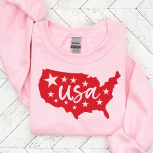 K&ouml;nnte beinhalten: Rosafarbener Pullover mit einer roten Umrandung der Vereinigten Staaten und dem Wort "usa" in wei&szlig;en Buchstaben innerhalb der Umrandung. Um die Umrandung herum befinden sich wei&szlig;e Sterne.