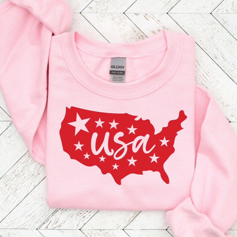 K&ouml;nnte beinhalten: Rosafarbener Pullover mit einer roten Umrandung der Vereinigten Staaten und dem Wort "usa" in wei&szlig;en Buchstaben innerhalb der Umrandung. Um die Umrandung herum befinden sich wei&szlig;e Sterne.