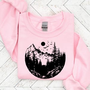 May include: Pink sweatshirt with a black graphic of a mountain range, trees, and a moon with birds flying above. The graphic is in a circle shape.