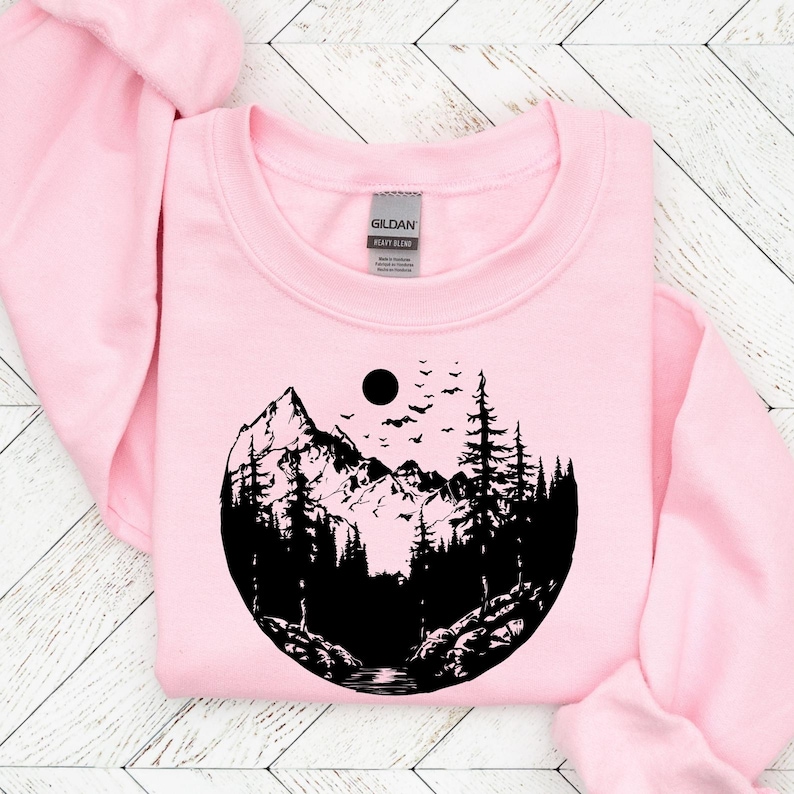 May include: Pink sweatshirt with a black graphic of a mountain range, trees, and a moon with birds flying above. The graphic is in a circle shape.