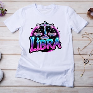 Op de afbeelding: Wit T-shirt met een ontwerp van het sterrenbeeld Weegschaal. De afbeelding toont een weegschaalsymbool met roze en blauwe verlopen, en het woord "LIBRA" in een gestileerd lettertype. Het ontwerp staat tegen een zwarte achtergrond.
