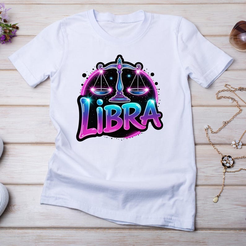 Op de afbeelding: Wit T-shirt met een ontwerp van het sterrenbeeld Weegschaal. De afbeelding toont een weegschaalsymbool met roze en blauwe verlopen, en het woord "LIBRA" in een gestileerd lettertype. Het ontwerp staat tegen een zwarte achtergrond.