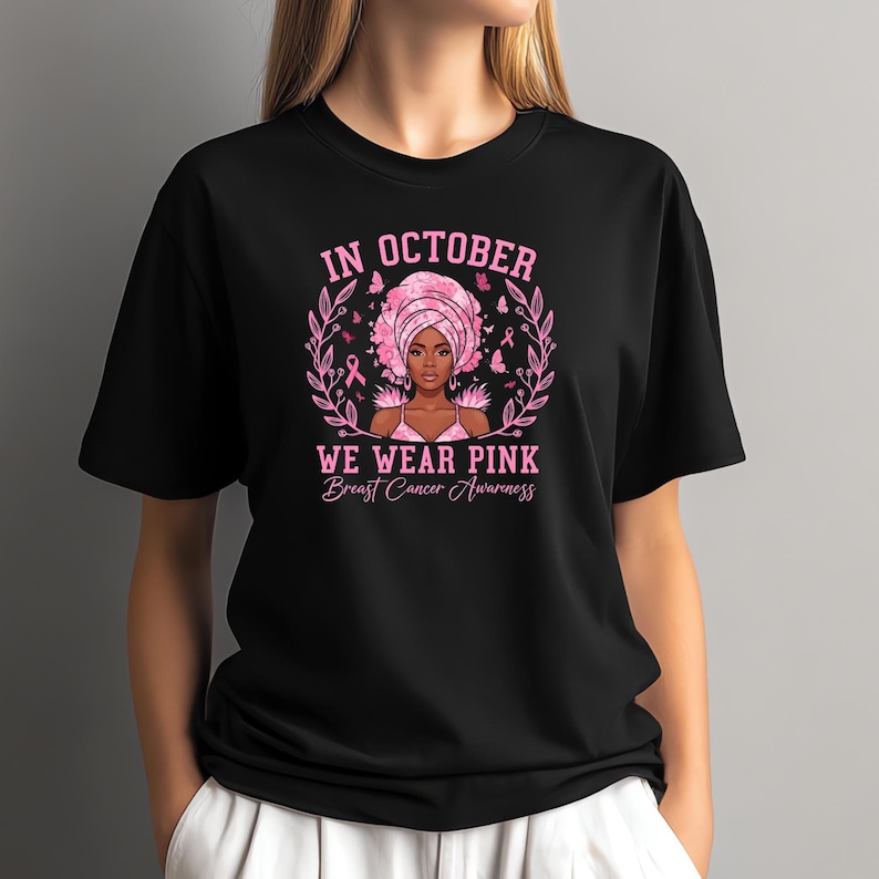 May include: Black t-shirt with a pink graphic for Breast Cancer Awareness. The graphic includes the text "IN OCTOBER WE WEAR PINK Breast Cancer Awareness" with an illustration of a woman wearing a pink head wrap.