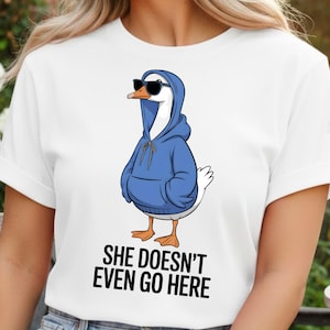 May include: White t-shirt featuring a cartoon goose wearing a blue hoodie and sunglasses. The goose has orange feet and a black beak. The text "SHE DOESN'T EVEN GO HERE" is printed in black below the goose.