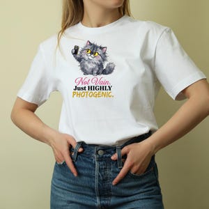 May include: A white short-sleeved t-shirt featuring a graphic of a fluffy grey cat taking a selfie with a smartphone. Below the cat, the text reads: Not Vain. Just HIGHLY PHOTOGENIC. The shirt is paired with dark blue denim jeans with a button fly.