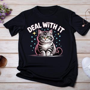 K&ouml;nnte beinhalten: Schwarzes T-Shirt mit einer Cartoon-Katze mit grau-wei&szlig;en Streifen, gr&uuml;nen Augen und dem Text "DEAL WITH IT" in Wei&szlig;. Das Design beinhaltet Sterne und blaue Punkte. Das Shirt wird auf einer hellen Holzoberfl&auml;che pr&auml;sentiert.